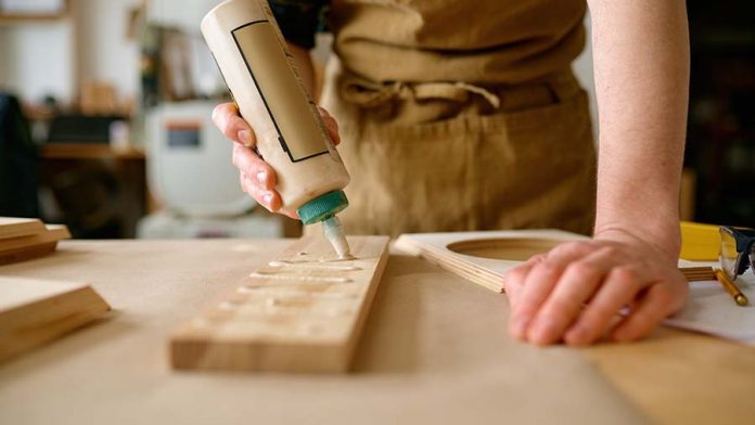 A carpenter in a brown work apron applies wood glue to a piece of pine board in a zig-zag pattern.