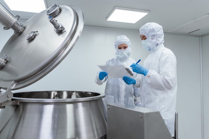Two workers in white cleanroom suits and blue gloves reviewing documentation next to an open stainless steel mixing vessel in a manufacturing facility.