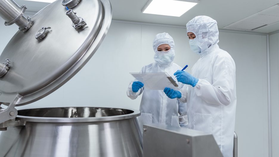 Two workers in white cleanroom suits and blue gloves reviewing documentation next to an open stainless steel mixing vessel in a manufacturing facility.